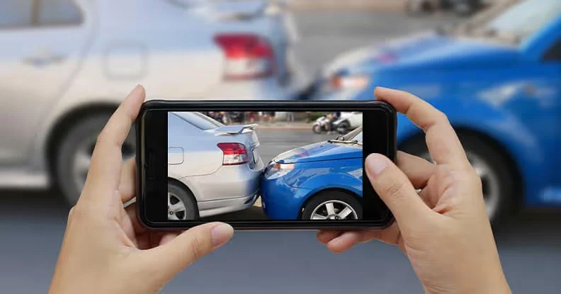 Hands holding a phone to photograph a collision between a silver car and a blue car
