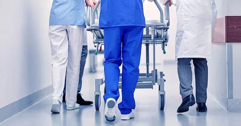 Hospital staff in scrubs and a white coat pushing a stretcher down a corridor