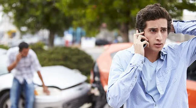 Young man on the phone after a car crash, with the damaged cars and the other driver behind him