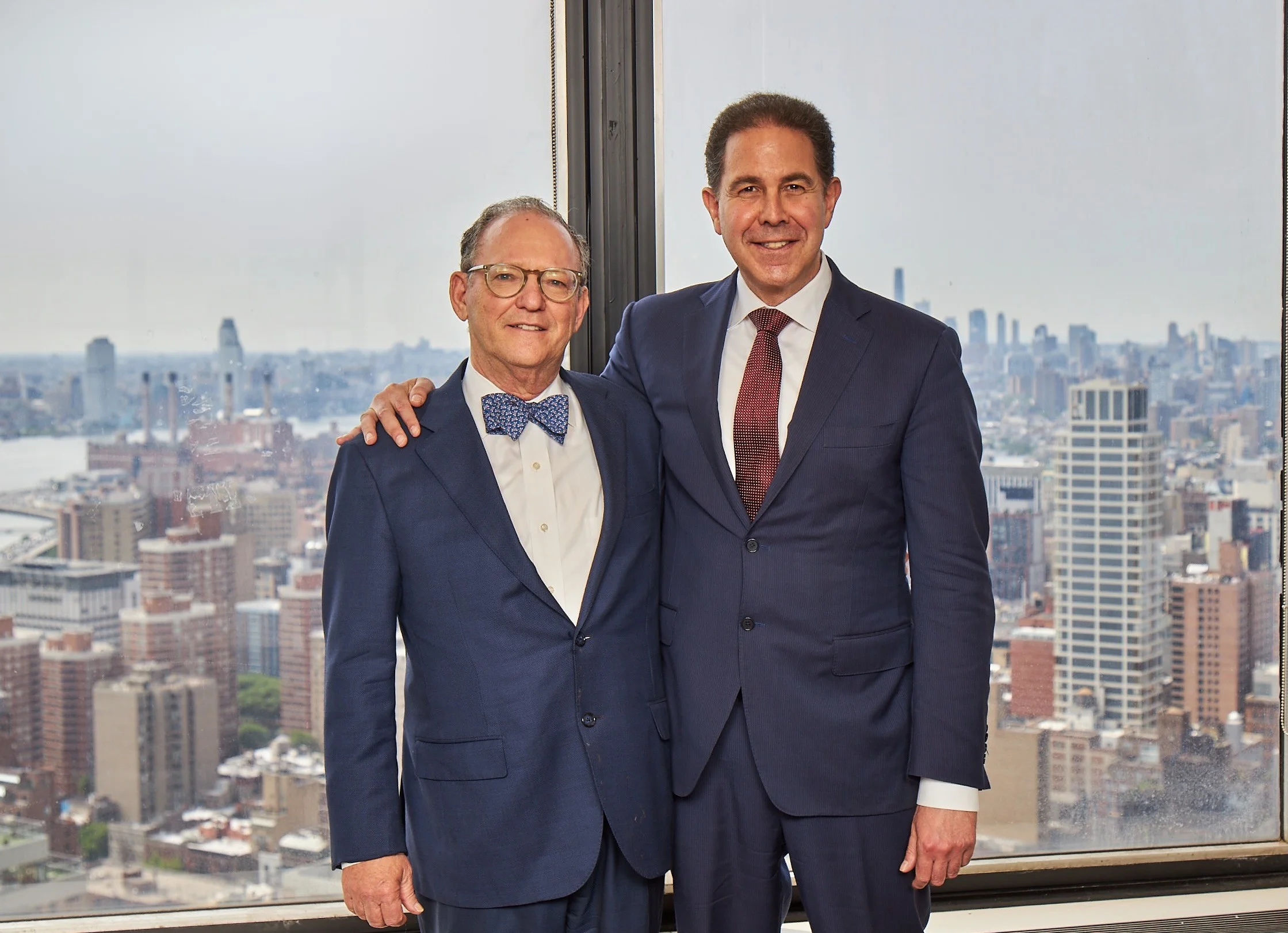 Attorneys Alan Fuchsberg and Bradley Zimmerman in suits standing by an office window overlooking Manhattan
